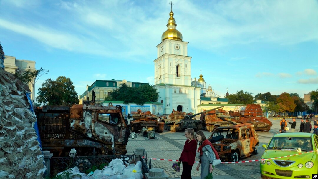 Two women holding bouquets of flowers stand in front of a display of destroyed Russian tanks and armored vehicles near St. Michael's Golden-Domed Monastery in Kyiv, Sept. 11, 2022