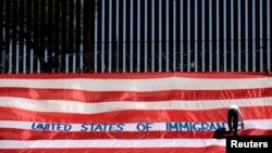 Roberto Marquez, yang dikenal sebagai Roberz, menulis pada bendera AS sebagai bagian aksi 'United States of Immigrants', yang memprotes perlakuan tak manuasiawi terhadap para migran, di tembok perbatasan di El Paso, Texas, 6 Juni 2019. 