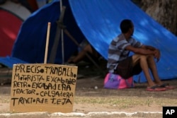 In this March 11, 2018 photo, a sign that reads in Portuguese: "I need work. Tractor driver. Excavator." near a Venezuelan migrant outside his tent in Simon Bolivar Square where Venezuelans have set up tents.