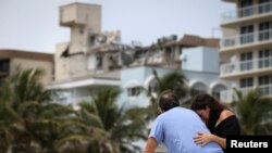 Sepasang suami istri di pantai bereaksi di dekat bangunan tempat tinggal yang sebagian runtuh ketika kru darurat melanjutkan operasi pencarian dan penyelamatan untuk para penyintas, di Surfside, dekat Miami Beach, Florida, AS 26 Juni 2021. (Foto: REUTERS/Marco Bello)