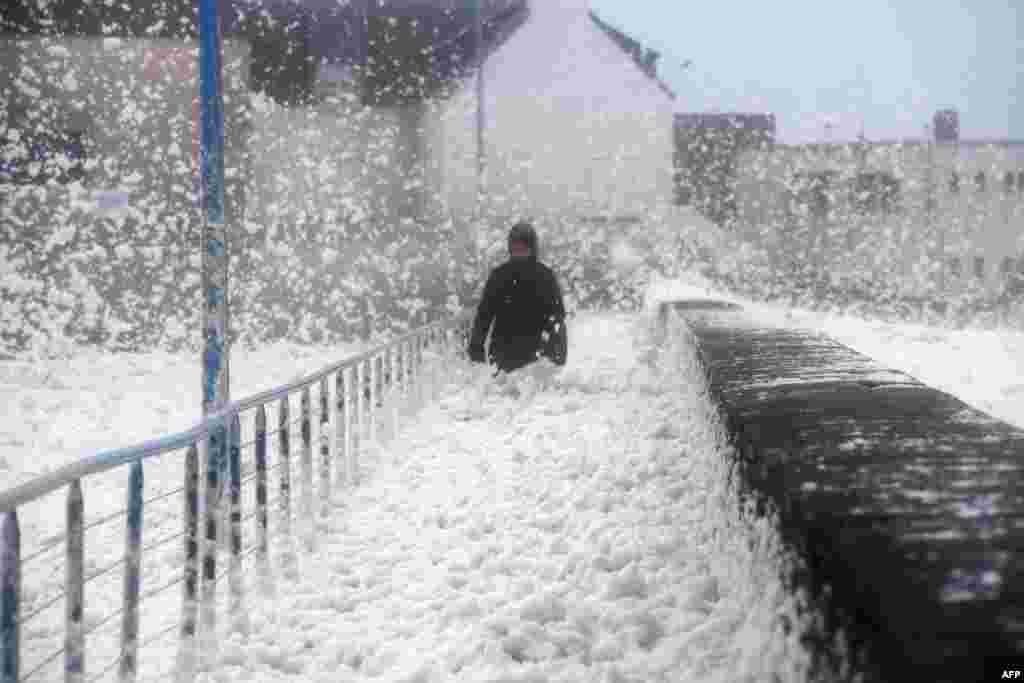 A person walks as sea foam washes up on the frontwalk in Saint-Guenole, western France, as storm Dennis sweeps accross Brittany.