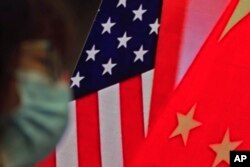 In this Feb. 22, 2021, file photo, a woman wearing a face mask sits near a screen showing China and U.S. flags as she listens to a speech on China-U.S. relations.