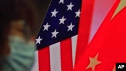 Seorang wanita mengenakan masker duduk di dekat layar yang menunjukkan bendera China dan AS saat dia mendengarkan pidato tentang hubungan kedua negara. 