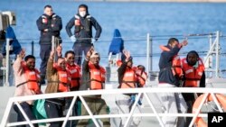 Para migran melambaikan tangan dan memberi tanda dua jari membentuk huruf v turun di Hay Wharf, Pieta, Malta, 9 Januari 2019. 