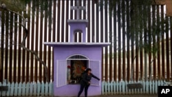En esta foto de enero 16 de 2019, Esther Arias abre una ventana en una capilla construida por su padre José Arias delante de una renovada sección de una valla fronteriza de acero en Tijuana, México. 