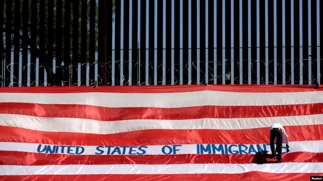 Roberto Marquez, yang dikenal sebagai Roberz, menulis pada bendera AS sebagai bagian aksi 'United States of Immigrants', yang memprotes perlakuan tak manuasiawi terhadap para migran, di tembok perbatasan di El Paso, Texas, 6 Juni 2019. 