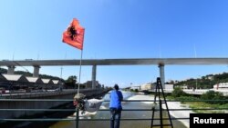 FILE - A man stands next to flowers and a t-shirt with the picture of a victim of the collapse of the Morandi bridge, as static testing operations are performed on the bridge ahead of its inauguration, in Genoa, Italy, July 19, 2020.