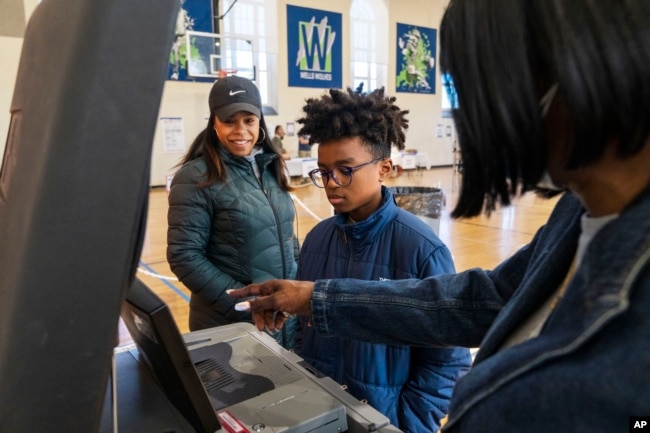 Zari Gedeon, 11, center, listens to a poll worker as he helps his mother, Lauren Gedeon, of Washington, left, submit her vote along with his siblings McKenzie, 8, and Brooklyn, 4, during voting in the midterm elections, Tuesday, Nov. 8, 2022, at Ida B. Wells Middle School in Washington. (AP Photo/Jacquelyn Martin)