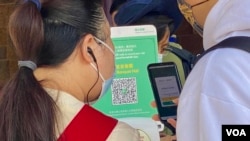 一名香港市民(白衣者)正准备使用防疫手机应用程式“安心出行”进入一间餐厅用膳，身旁的餐厅职员检视使用者是否有准确扫瞄餐厅入口的二维码 (美国之音/汤惠芸)