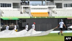 ডোমিনিকার রোসেউর উইন্ডসর পার্কে ওয়েস্ট ইন্ডিজ এবং বাংলাদেশের মধ্যে প্রথম টি-টোয়েন্টি খেলা চলাকালীন বৃষ্টি শুরু হওয়ায় মাঠের কর্মীরা পিচ ঢাকছেন। (ছবি র‍্যান্ডি ব্রুকস/এএফপি)