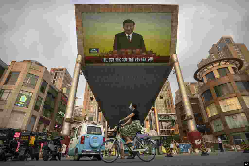 A woman wearing a face covering rides a bicycle past a large television screen at a Beijing shopping center showing Chinese state television news coverage of Chinese President Xi Jinping&#39;s visit to Hong Kong.