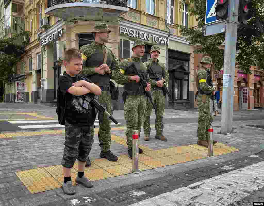 Seorang anak laki-laki dengan senapan mesin mainan berdiri di dekat empat tentara Ukraina saat mereka berpatroli di sebuah daerah di ibu kota Kyiv, di tengah serangan Rusia di Ukraina. (Foto: Reuters)&nbsp;