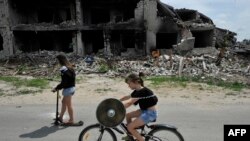 Children ride a bike and a scooter on a road in front of a destroyed building in the village of Novoselivka, outside Chernigiv, on June 21, 2022.