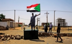 FILE - A Sudanese protester holds a national flag as he stands on a barricade along a street, demanding that the country's Transitional Military Council hand over power to civilians, in Khartoum, Sudan, June 5, 2019.