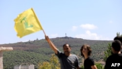 A man waves the Hezbollah flag in the southern Lebanese village of Aita al-Shaab, which has come under Israeli fire numerous times, June 29, 2024. An Israeli military position can be seen in the background.