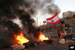 FILE - An anti-government protester carries her national flag past burning tires blocking the main highway during protests against the corruption in Khaldeh, south of Beirut, Lebanon, Nov. 13, 2019.
