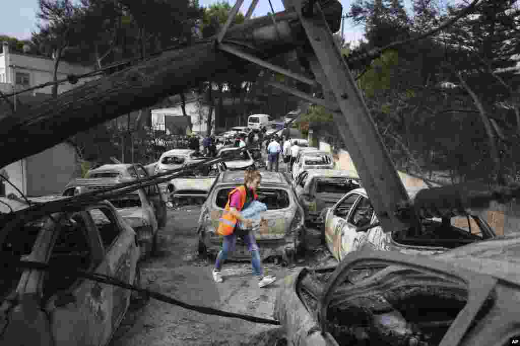 A woman carries bottles of water as people stand amid the charred remains of burned-out cars in Mati east of Athens, Tuesday, July 24, 2018.