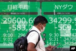 A man wearing a protective mask walks in front of an electronic stock board showing Japan's Nikkei 225 and New York Dow indexes at a securities firm, Sept. 29, 2021, in Tokyo.