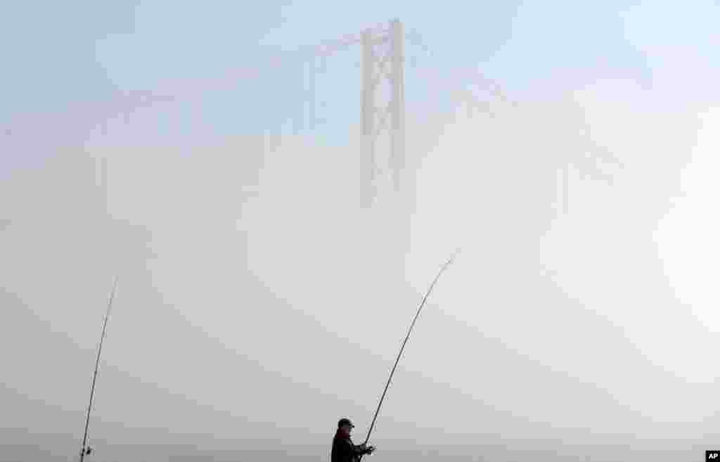Seorang memancing ikan di sungai Tagus sementara kabut tebal menyelimuti jembatan di Lisabon, Portugal.