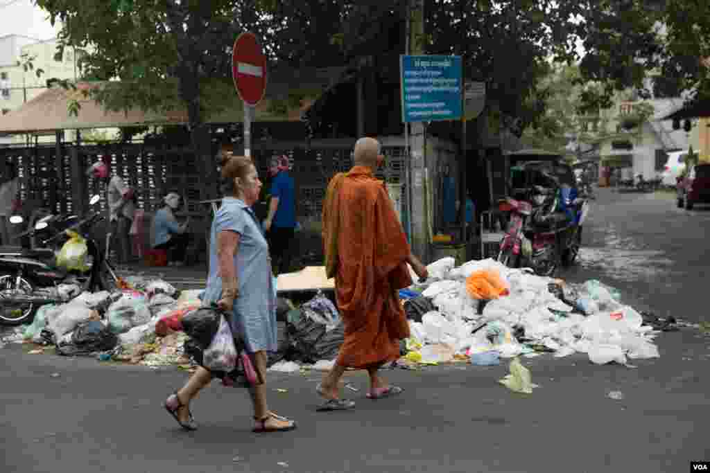 ស្រ្តីម្នាក់និងព្រះសង្ឃមួយអង្គដើរកាត់គំនរសម្រាមនៅក្នុងរាជធានីភ្នំពេញមួយថ្ងៃបន្ទាប់ពីបុគ្គលិកក្រុមហ៊ុនស៊ីនទ្រីមិនមកប្រមូលក្នុងបាតុកម្មទាមទារតម្លើងប្រាក់បៀវត្សកាលពីថ្ងៃទី២៦ ខែធ្នូ ឆ្នាំ២០១៨ ប្រទេសកម្ពុជា។(ទុំ ម្លិះ/VOA)