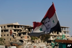 In this Sunday, July 15, 2018, photo, a Syrian national flag with the picture of President Bashar Assad hangs at an Army check point, in the town of Douma in the eastern Ghouta region, near the Syrian capital Damascus, Syria.