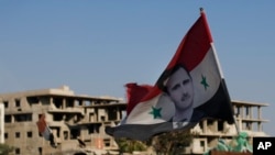 In this Sunday, July 15, 2018, photo, a Syrian national flag with the picture of President Bashar Assad hangs at an Army check point, in the town of Douma in the eastern Ghouta region, near the Syrian capital Damascus, Syria.