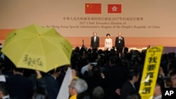 Protesters raises an umbrella and placard reading: "I want genuine universal suffrage" to protest as former Hong Kong Chief Secretary Carrie Lam, center, declares her victory in the chief executive election of Hong Kong while former Financial Secretary John Tsang, left, and retired judge Woo Kwok-hing stand with her in Hong Kong, March 26, 2017. 