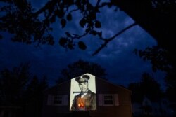 An image of veteran Samuel Melendez is projected onto the home of his nieces, Janet Ramirez, right, and Mary Perez, as they look out a doorway in Chicopee, Mass., May 17, 2020.