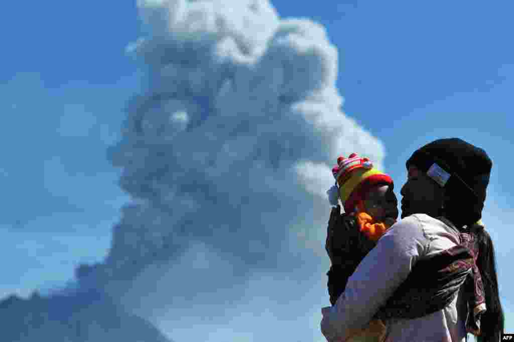A woman carries her child as she watches Mount Sinabung volcano spewing thick volcanic ash, as seen from Karo, Indonesia.