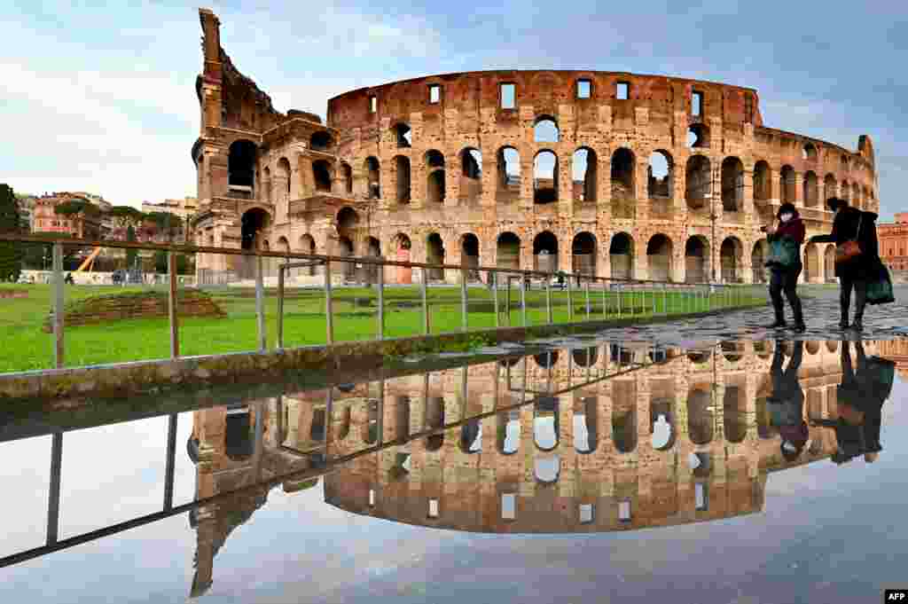 People walk past the Colosseum monument in Rome, reflected in a pool of water following heavy rains.