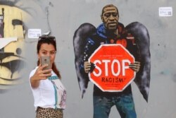 A woman takes a selfie picture posing next to a street poster artwork by Italian urban artist Salvatore Benintende aka "TVBOY" depicting George Floyd with angel wings and holding a stop traffic sign against racism, in a street of Barcelona