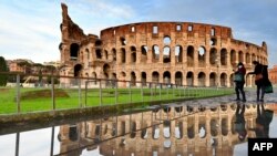 People walk past the Colosseum monument in Rome, reflected in a pool of water following heavy rains.