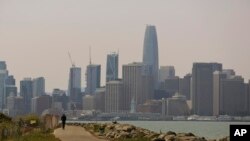 A man rides a scooter on Treasure Island as the San Francisco city skyline sits in a smoky haze in the background Wednesday, Aug. 8, 2018. 