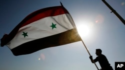 FILE - A Syrian protester stands on top of a building as he waves a Syrian flag, during a demonstration against a U.S. raid at a village near the Syrian-Iraqi border, in Damascus, Syria, Oct. 30, 2008. Hundreds of Syrian riot police ringed the shuttered and closed U.S. Embassy in Damascus as tens of thousands of Syrians converged on a central square for a government-orchestrated protest to denounce a deadly U.S. raid near the Iraqi border.