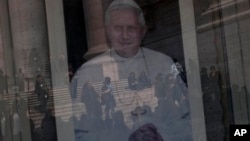 Tourists are reflected in a portrait of Pope Benedict XVI's in St. Peter's Square, a day before the Pope's last general audience, the Vatican, Feb. 26, 2013.