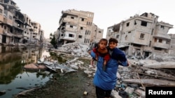 Një palestinez mban në krahë fëmijët e tij ndërsa ecën pranë ndërtesave të shkatërruara nga sulmet izraelite në Khan Younis
