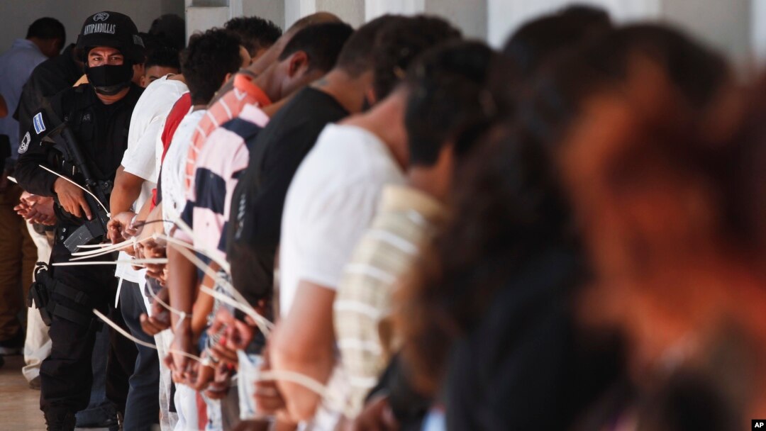 FILE - A policeman of an anti-gang unit stands next to handcuffed alleged members of the Mara Salvatrucha (MS-13) detained during a police raid in San Salvador, El Salvador, Jan. 31, 2014.