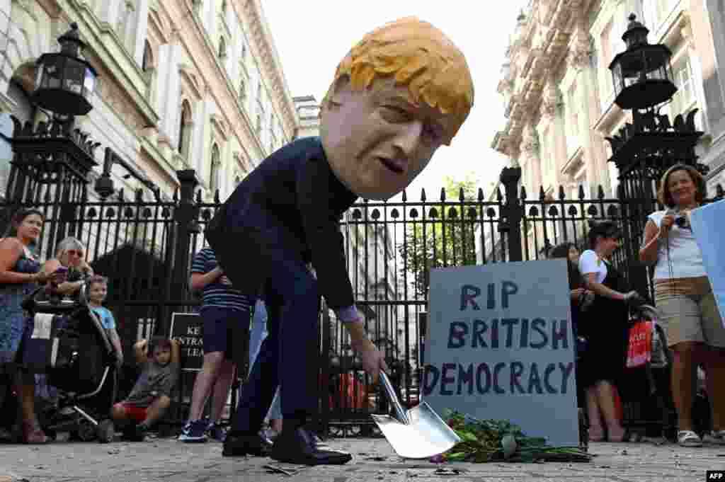 A demonstrator wearing a mask depicting Britain&#39;s Prime Minister Boris Johnson stands beside a mock gravestone inscribed with the words &quot;RIP British Democracy&quot; outside the gates to Downing Street in central London.