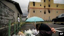 In this July 19, 2019, file photo, a woman prays at a makeshift memorial to honor the victims of a fire at the building of Kyoto Animation's No. 1 studio, background, in Kyoto, western Japan. 