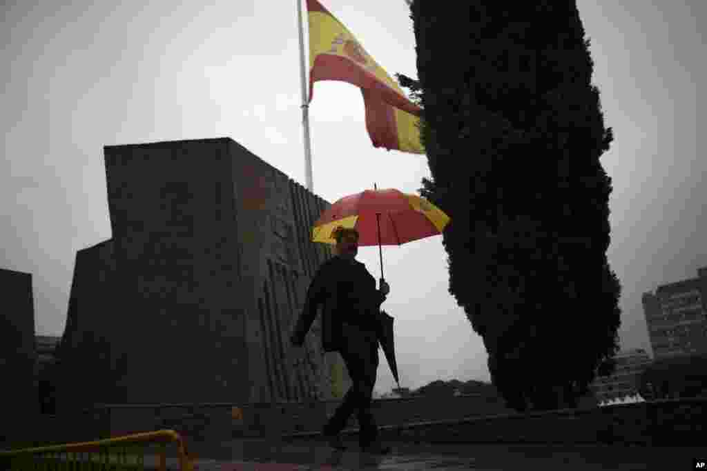 مردی در زیر باران با چتری به شکل پرچم اسپانیا در حال قدم زدن در میدان کلمبوس مادرید اسپانیا