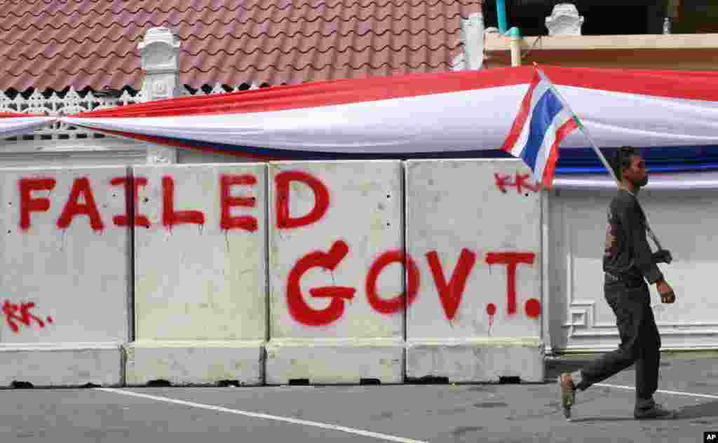 A protester with a Thai national flag walks past concrete barriers sprayed with "Failed Government" outside the fence around Government House, wrapped by a long banner in the colors of the national flag in Bangkok, Dec. 13, 2013. 