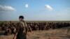 FILE - U.S.-backed Syrian Democratic Forces fighters stand guard next to men waiting to be screened after being evacuated out of the last territory held by Islamic State group militants, near Baghouz, eastern Syria, Feb. 22, 2019.