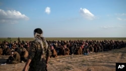 FILE - U.S.-backed Syrian Democratic Forces fighters stand guard next to men waiting to be screened after being evacuated out of the last territory held by Islamic State group militants, near Baghouz, eastern Syria, Feb. 22, 2019.