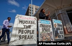 Pedestrians check their mobiles as they walk past a newspaper stand on a street in downtown Harare, Zimbabwe Sunday, Nov. 19, 2017. Zimbabwe's ruling party Central Committee says longtime President Robert Mugabe must resign as president by noon Monday.