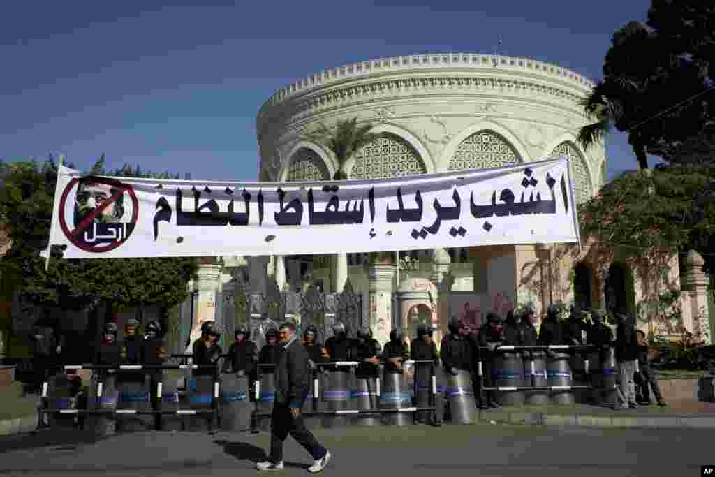 An Egyptian passes riot policemen guarding a gate of the presidential palace under a banner with a defaced picture of president Mohammed Morsi and Arabic that reads "the people want to bring down the regime," at the protests site, in Cairo, Egypt, Saturda
