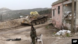 FILE - Syrian troops stand, with a destroyed tank in the background, on a street in Salma, Syria, Jan. 22, 2016. Syrian government forces, relying on Russian air cover, have recently seized Salma, located in Syria's province of Latakia, from militants.