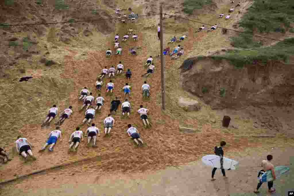 Two surfers walk toward the Mediterranean Sea as Israeli high-school seniors preparing to join the Israeli military later this year crawl out in sand dunes during an exercise at a privately run training camp for military combat fitness, in Herzliya, Israel, Feb. 1, 2019.