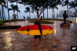 A surfer walks along Waikiki Beach in a light rain from Tropical Storm Lane, Aug. 25, 2018, in Honolulu.