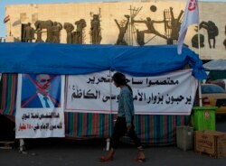 FILE - An anti-government protester walks by a defaced picture of Prime Minister-designate Adnan Al-Zurfi in Tahrir Square, Baghdad, Iraq, March 19, 2020.