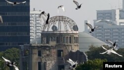 ພວກນົກກາງແກ ບິນຢູ່ເທິງສວນອະນຸສອນສັນຕິພາບ ຫຼື Peace Memorial Park ພ້ອມດ້ວຍຫໍ Atomic Bomb Dome ຢູ່ດ້ານຫຼັງ, ໃນພິທີລະນຶກເຖິງ ການຖິ້ມລະເບີດ ປາຣະມະນູໃສ່ນະຄອນ Hiroshima, ປະເ່ທດຍີ່ປຸ່ນ, ຮູບຖ່າຍ ວັນທີ 6 ສິງຫາ 2015. 